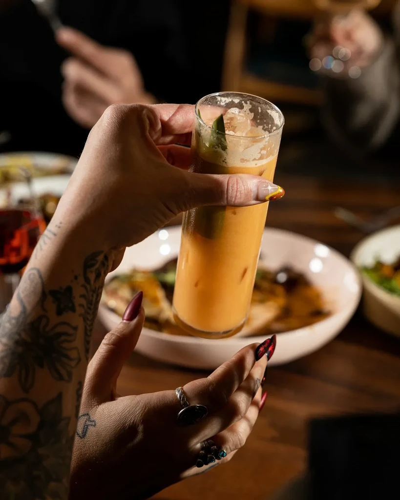 Tattooed hands with painted nails hold an iced amber drink.