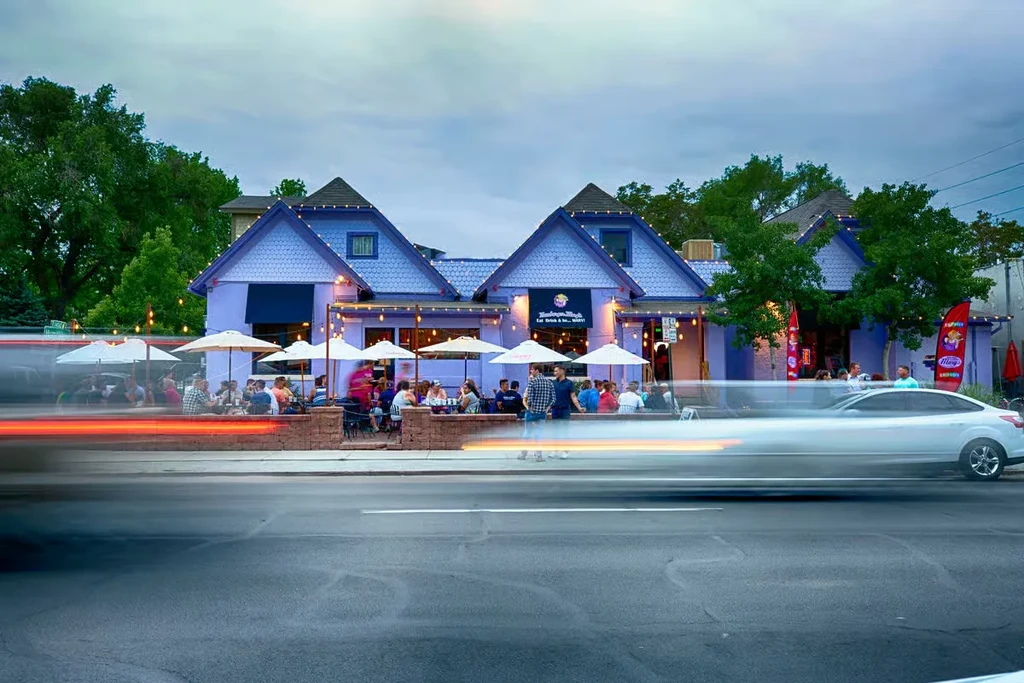 Purple restaurant with string-lit patio, patrons, blurred street at dusk.