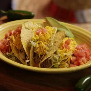 Four hard-shell tacos with meat, lettuce, cheese, tomatoes, and avocado.