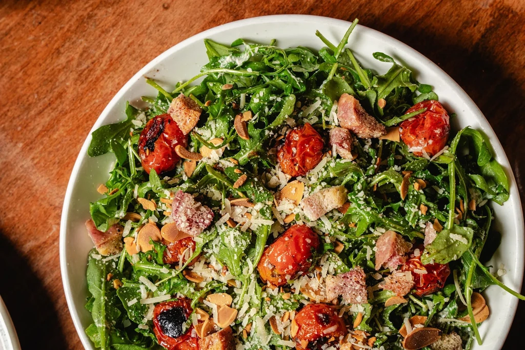 Overhead arugula salad with roasted tomatoes, shaved cheese, almonds, bacon.