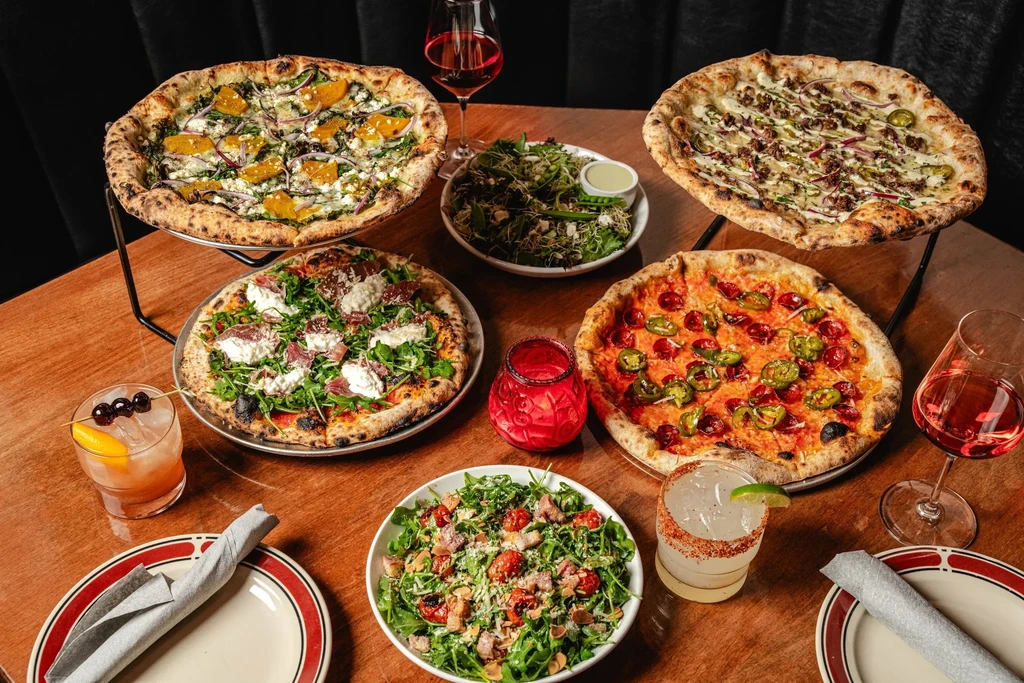 Four distinct pizzas, two salads, and various drinks on a wooden table.