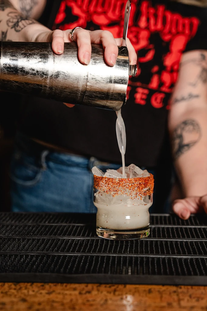 Bartender's tattooed hands pour a chili-rimmed cocktail into an icy glass.
