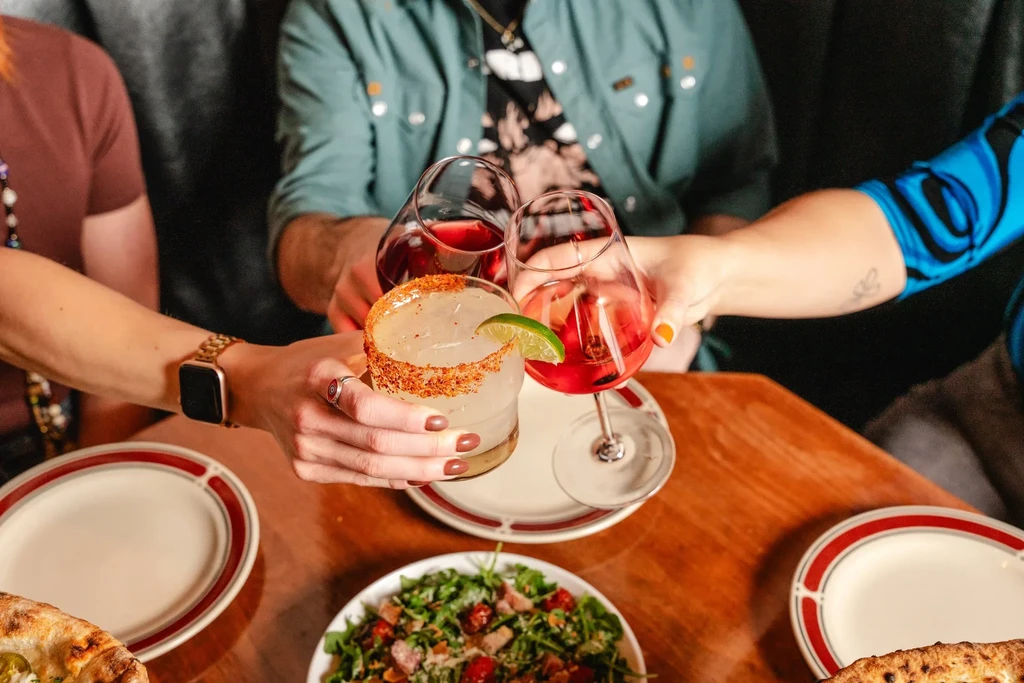 Three hands clink cocktails and wine over a table with pizza and salad.