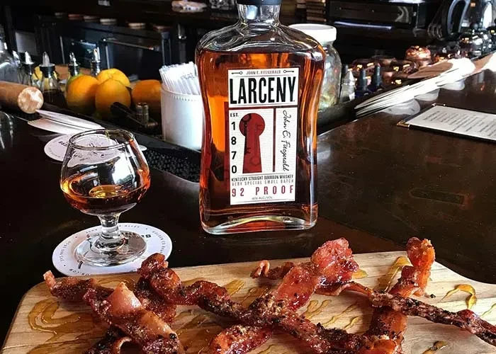Larceny bourbon, snifter, and candied bacon on a bar counter.