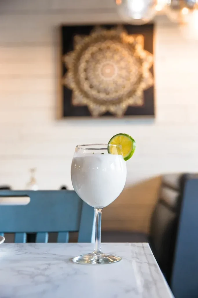 White beverage in stemmed wine glass with lime wedge on table.