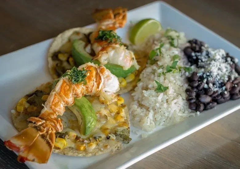 White plate with two lobster tacos, rice, black beans, and lime.