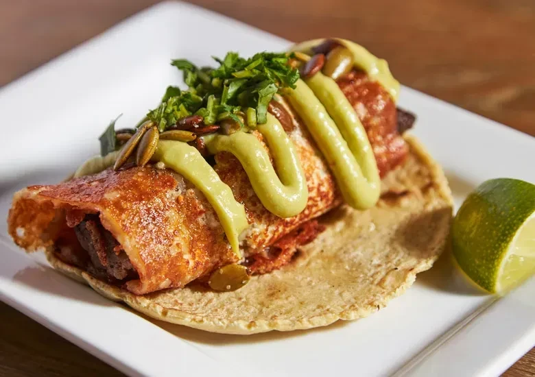 Crispy shelled taco with green sauce, herbs, pepitas, and a lime wedge.