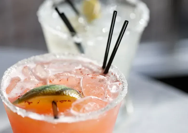 Two iced cocktails, one pinkish-orange with lime and one lighter.