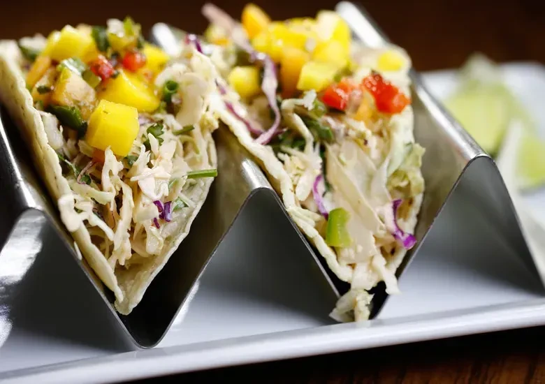 Two tacos in a silver holder with slaw, mango salsa, and lime.