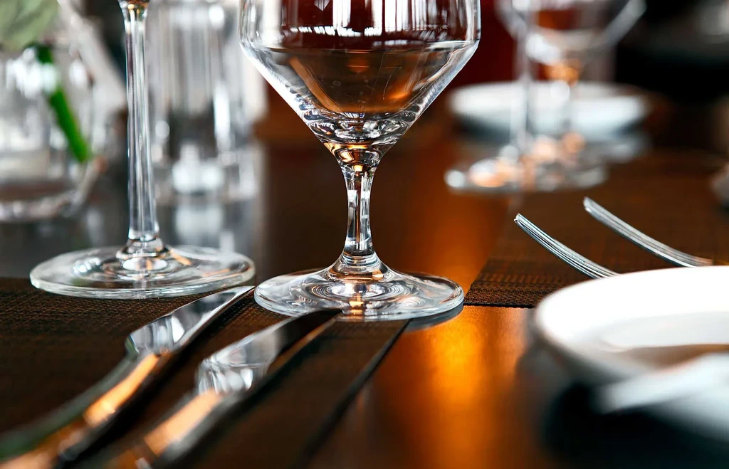 Warm-lit close-up of wine glass, silverware, and plate on elegant table.