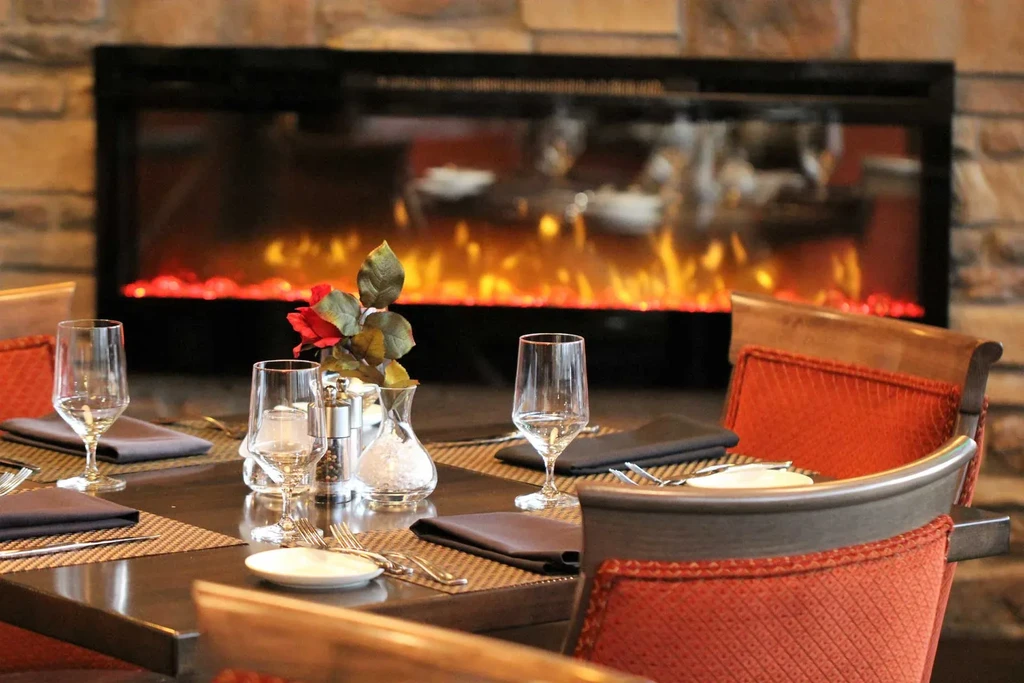 Warmly lit restaurant table with red flowers and electric fireplace.
