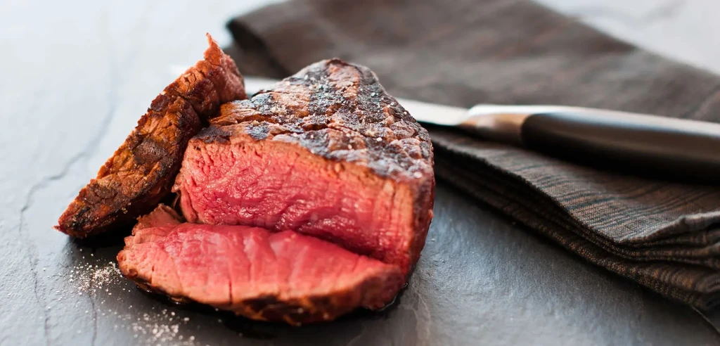 Seared medium-rare steak, cut open, with salt on a dark surface.