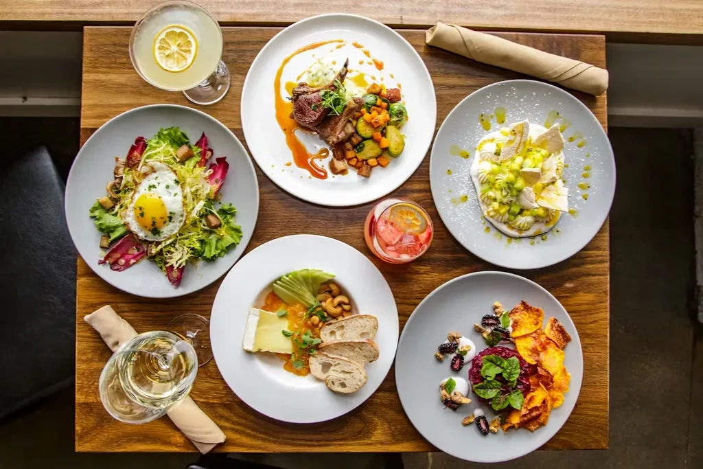 Overhead flat lay of six diverse prepared dishes and drinks on wood.