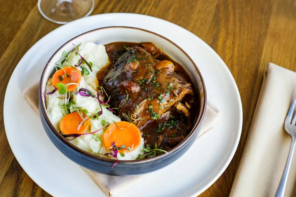 Plated braised short ribs, mashed potatoes, and carrots in a dark bowl.