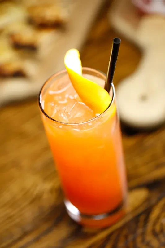 Vibrant orange-red drink with ice, spiral peel, and straw on wood.