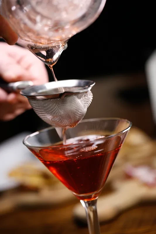 A bartender's hand straining vibrant red cocktail into a martini glass.