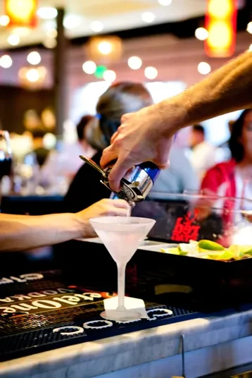 Hands pour clear liquid from shaker into frosted martini glass.