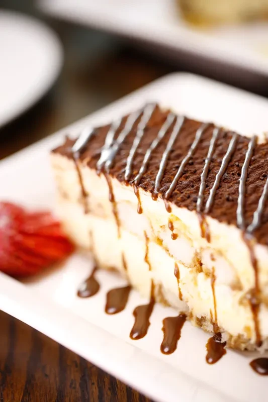 Tiramisu slice with cocoa, chocolate drizzle, and strawberry on a white plate.