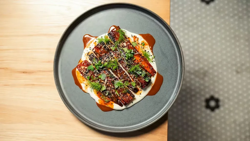 Glazed meat strips on creamy white sauce with microgreens and seeds.
