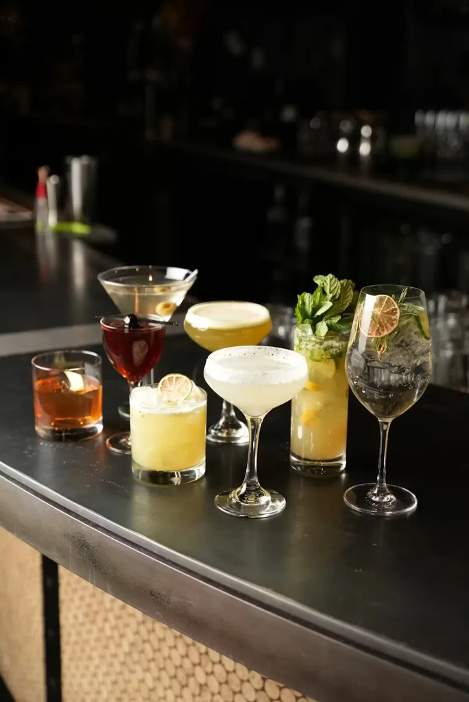 Seven garnished cocktails on a dark, reflective bar counter.
