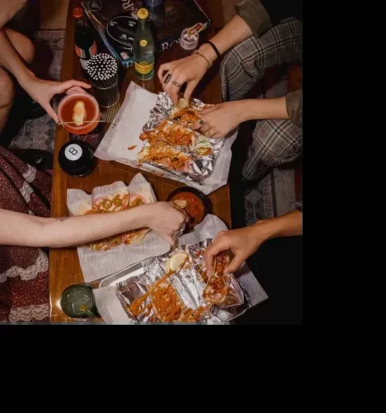 Hands share tacos, drinks, and an 8-ball on a wooden table.