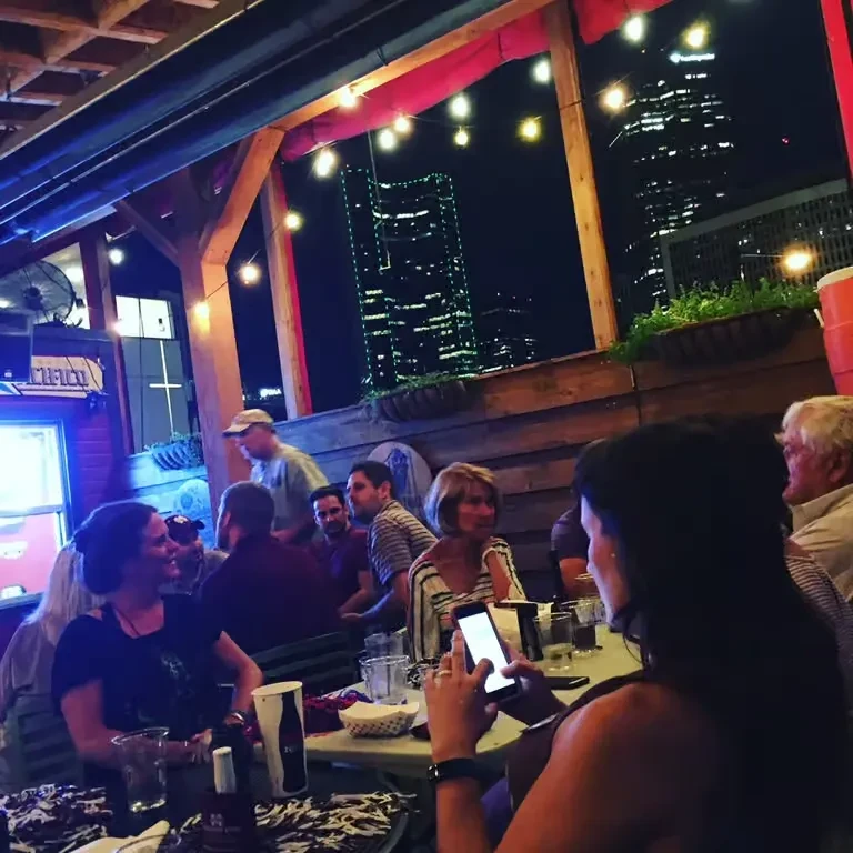 People socialize on a warm string-lit patio overlooking a city skyline.