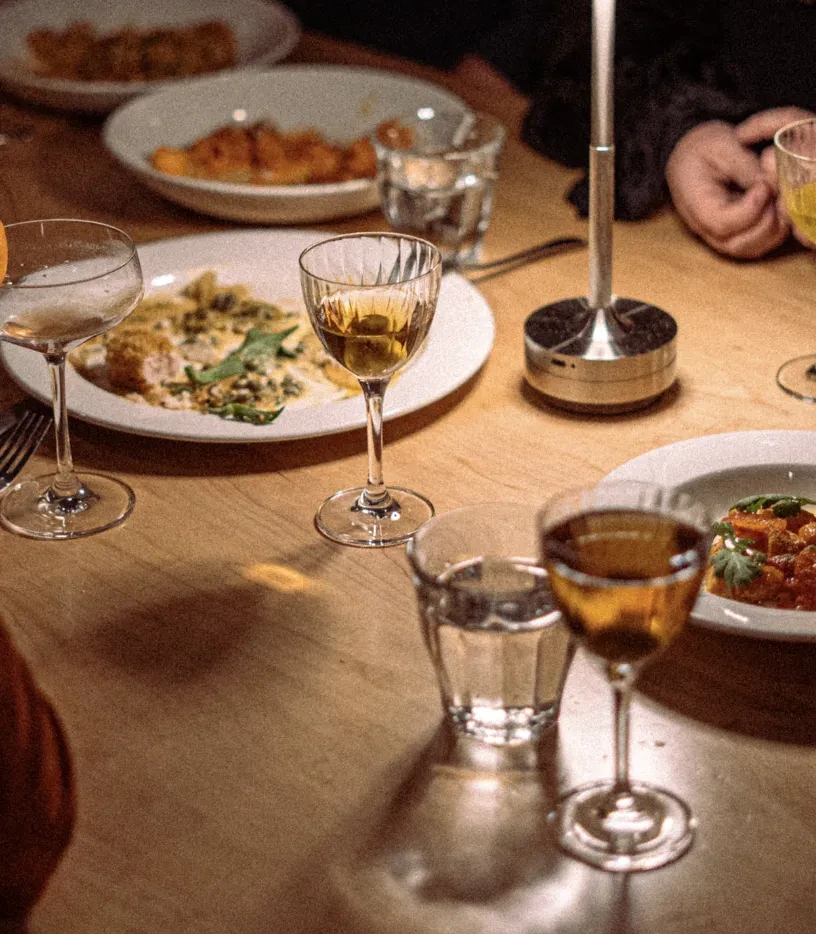 Warm, intimate dining table with multiple dishes, drinks, and diners' hands.