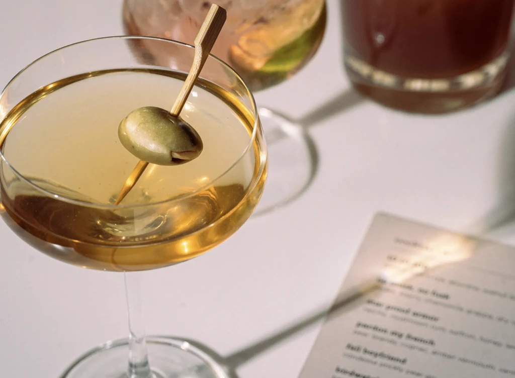 Golden-amber cocktail with green olive in a clear martini glass.