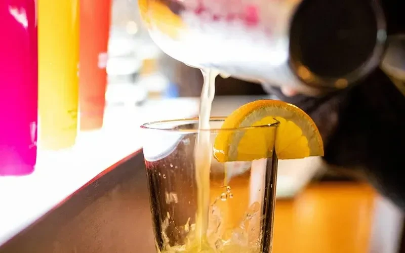 Light yellow cocktail poured from shaker into glass with orange slice.