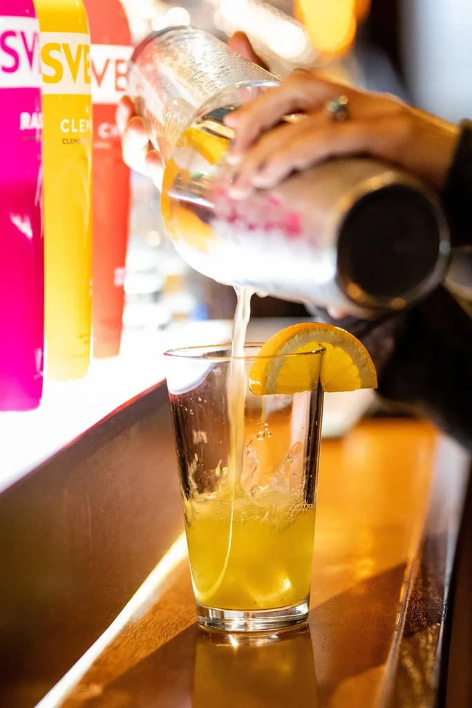 Bartender pouring yellow drink from shaker into iced glass with orange.