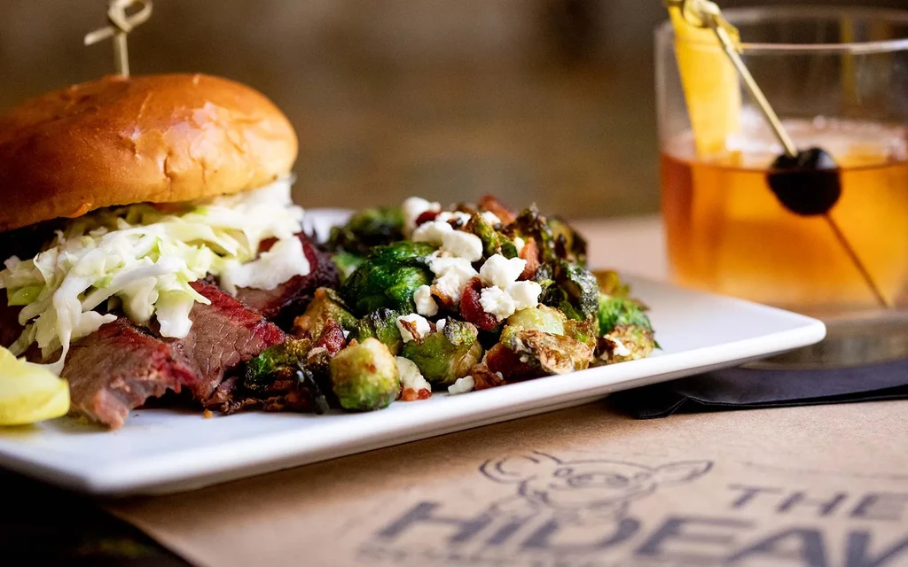 Large sandwich with coleslaw, roasted brussels sprouts, and an amber cocktail.