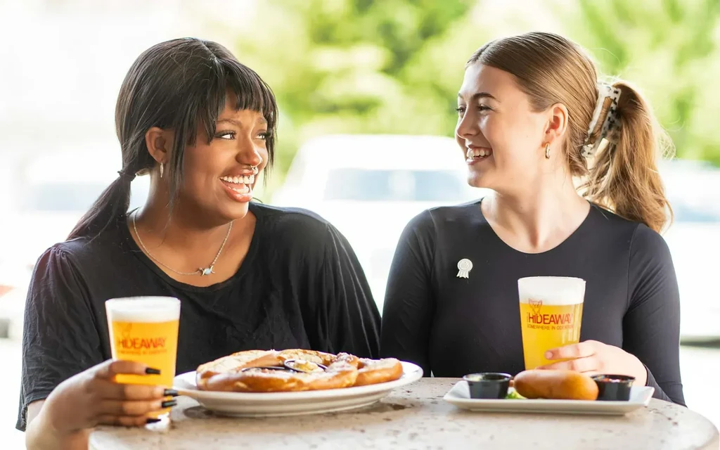 Two women cheerfully enjoying a pretzel, fried item, and beer.