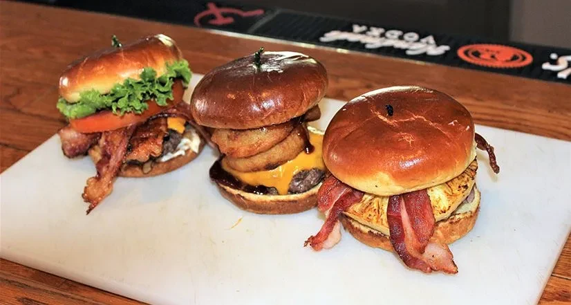 Three gourmet burgers with bacon, onion rings, and pineapple on a cutting board.