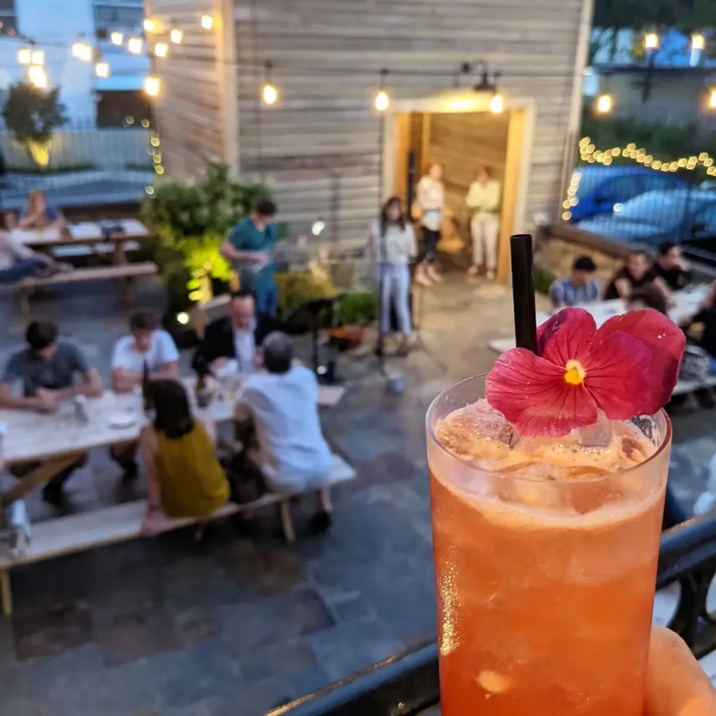 Reddish-orange cocktail with flower and straw, blurred lively evening patio.