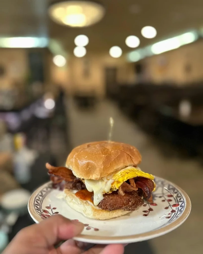 Hand holds breakfast sandwich with egg, cheese, bacon, hash brown.