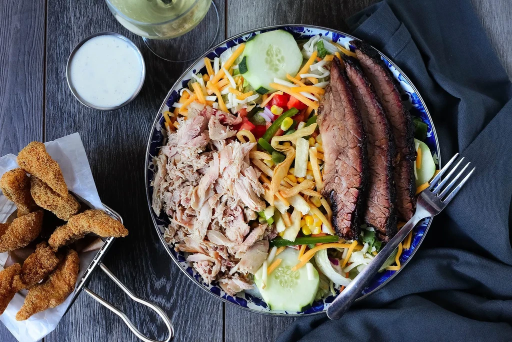 Large salad with meat, fried food, and sauce on a dark wooden table.