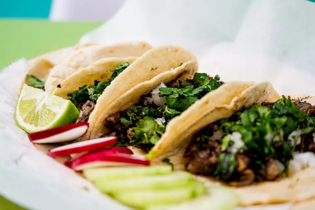 Three meat tacos with cilantro, onion, lime, radish, cucumber.