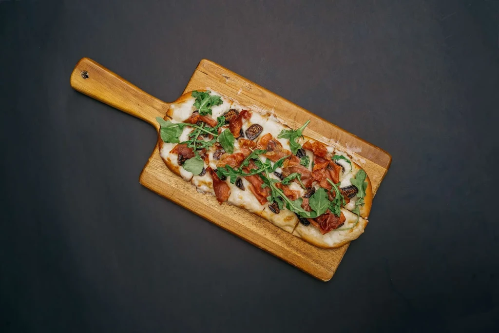 Rectangular flatbread with arugula, prosciutto, and dark figs on a wooden board.