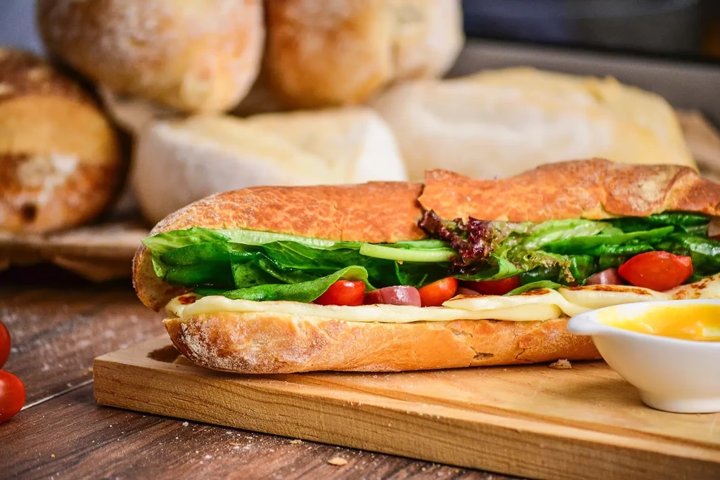 Baguette sandwich with lettuce, tomatoes, cheese, and red onions on a board.