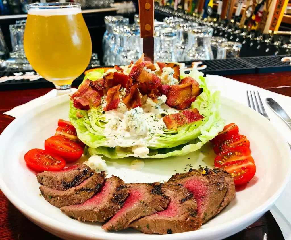 Plated medium-rare steak, wedge salad with knife, and beer at a bar.