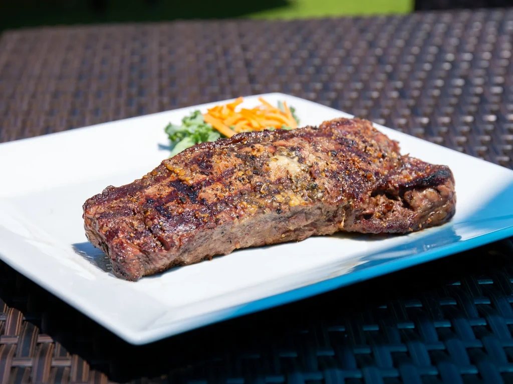 Well-seasoned grilled steak on a white plate with garnish, outdoors.