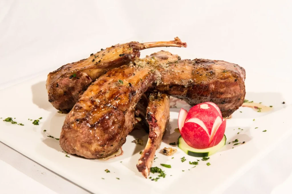 Glistening lamb chops with herbs, radish flower, and cucumber on white plate.