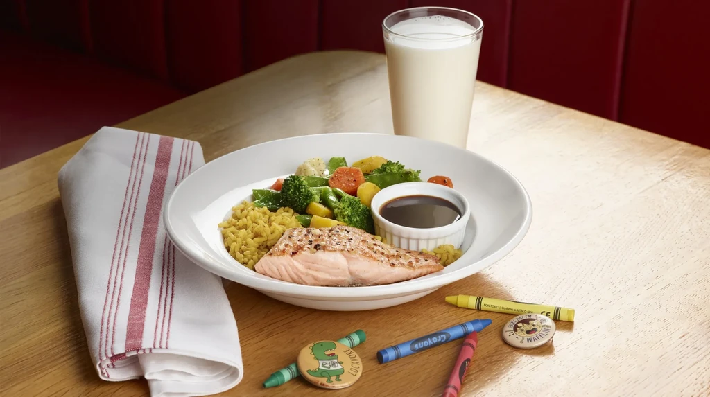 Grilled salmon meal, milk, crayons, and pins on restaurant table.