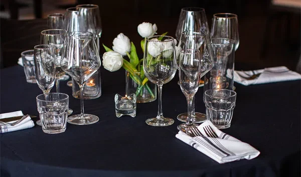 Set dining table with empty glasses, napkins, silverware, white flowers, lit candles.