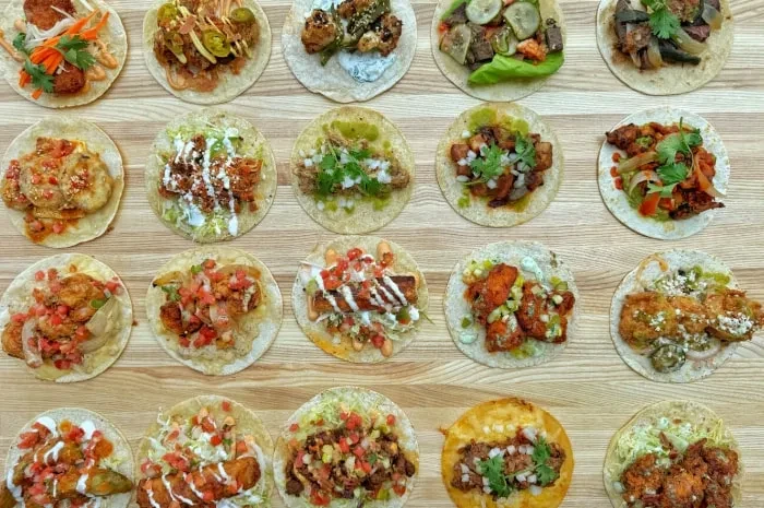Overhead view of 25 distinct, colorful tacos arranged on wood.