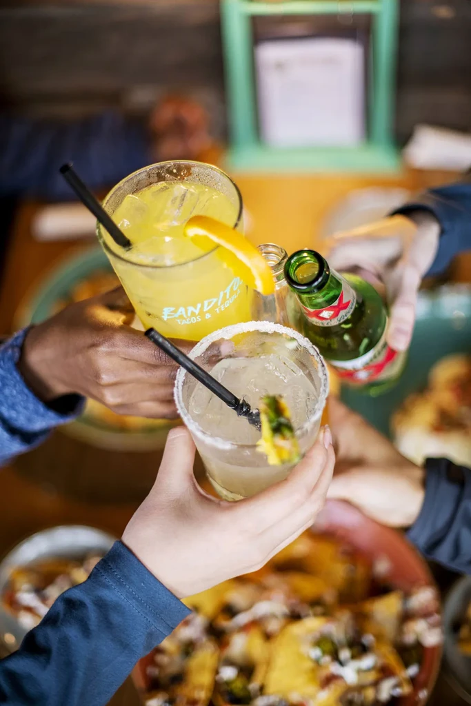Four hands clinking drinks, including a margarita and beer, at a restaurant.
