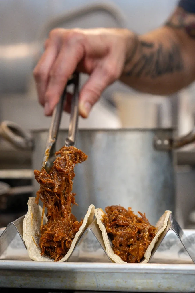 Hand filling a corn tortilla taco with shredded meat using tongs.