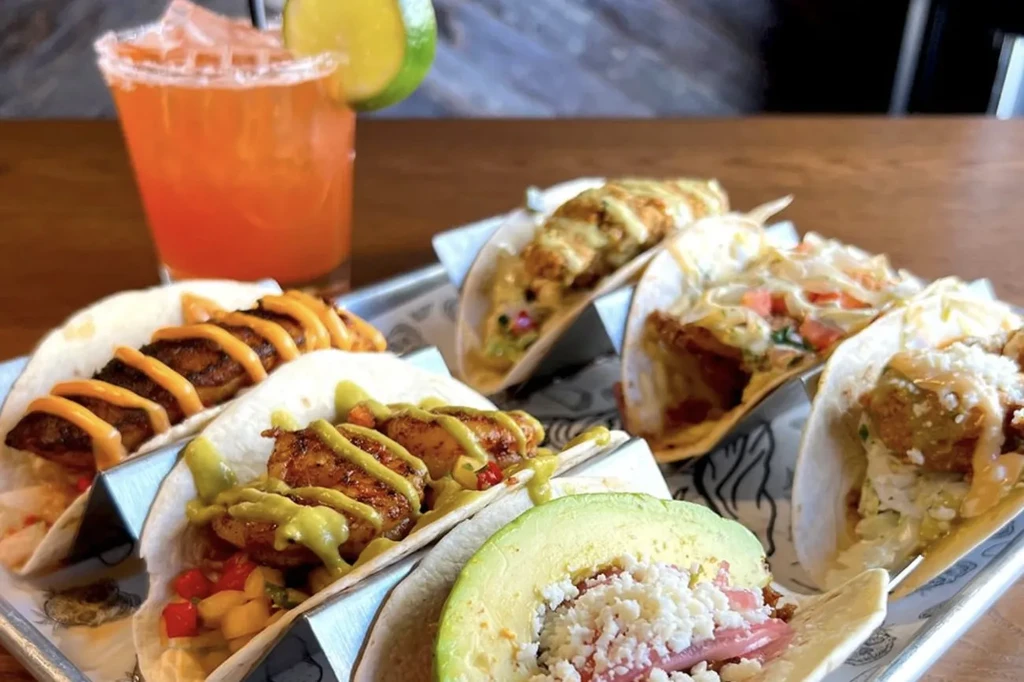 Metallic tray with various tacos and a cocktail on a wooden table.