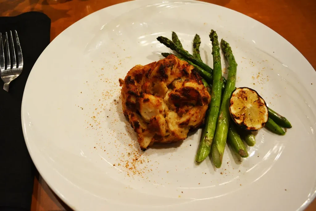 Golden-brown crab cake, asparagus, charred lemon, and fork on plate.
