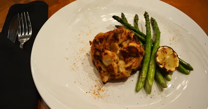 Plated seafood patty, asparagus, charred lemon, and seasoning on a white dish.